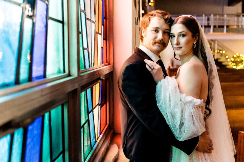A couple in wedding attire poses closely by a multicolored stained glass window, with soft lighting illuminating their faces.