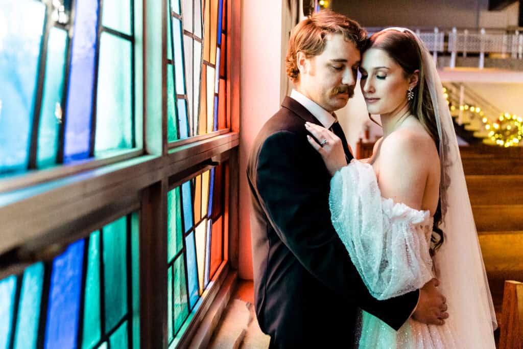 A bride and groom stand closely together, with their eyes closed, next to a stained glass window in a church. The groom is in a black suit and the bride is wearing a white gown and veil.