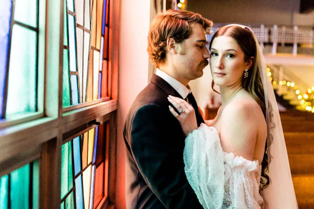 A couple in wedding attire stands by a colorful stained-glass window. The bride touches the groom's chest and looks towards the camera, while the groom gazes at her.