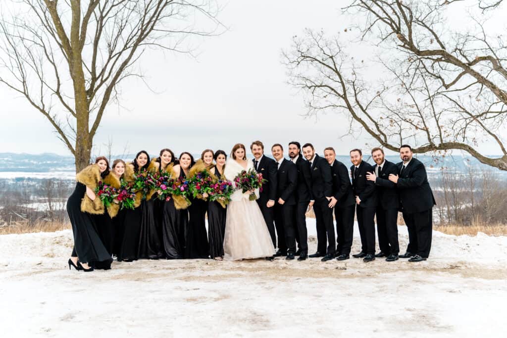 A large wedding party stands outdoors in winter attire, with the bride in the center holding a bouquet of flowers. Snow and bare trees are visible in the background.