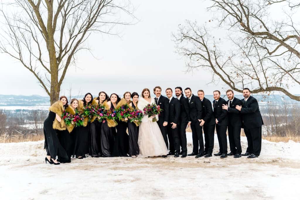 A wedding party poses outdoors in the snow, with the bride and groom at the center surrounded by bridesmaids in black dresses and fur stoles and groomsmen in black suits.