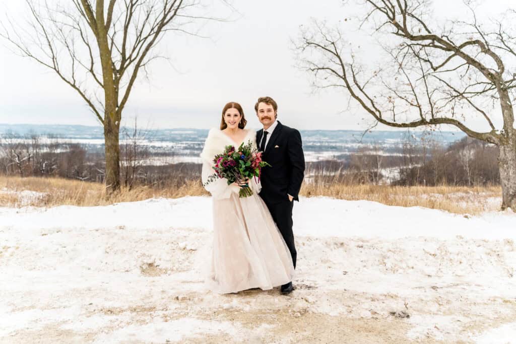 A bride and groom stand outdoors in a snowy landscape, with bare trees and distant hills in the background. The bride holds a bouquet of flowers.