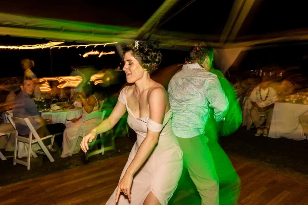 A woman in a light dress and a man in a shirt dance energetically on a wooden floor at a nighttime event with blurred lights and seated guests in the background.