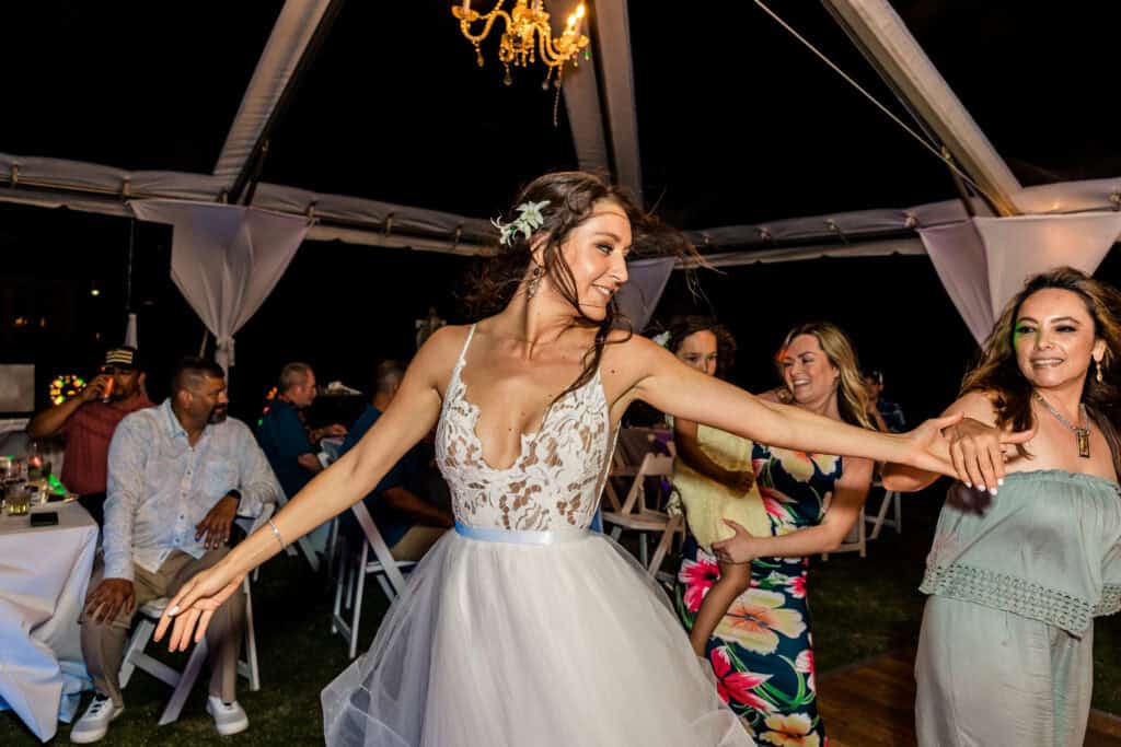 A bride in a white dress dances joyfully under a tent at night, holding hands with guests who are also dancing. A chandelier hangs from the tent ceiling, and other guests are seated in the background.