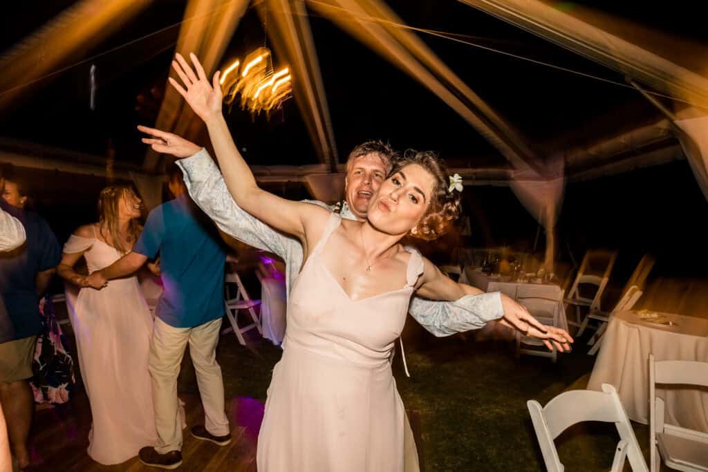 A couple is dancing enthusiastically at a party under a tent, with blurred lights and other attendees in the background. The woman has her arms outstretched, and the man stands closely behind her.