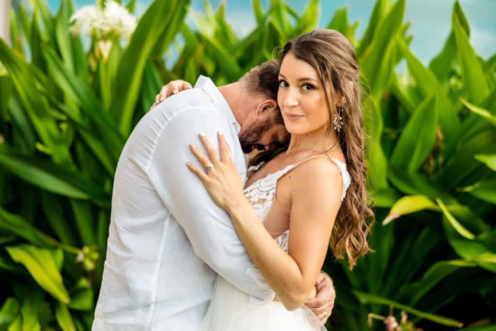 A woman in a white dress with a visible ring embraces a man in a white shirt while standing in front of lush tropical greenery.