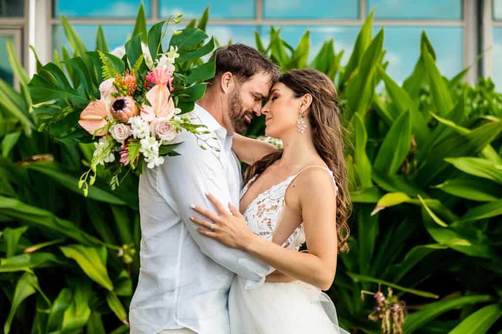 A couple in white attire embraces in front of lush greenery. The woman holds a bouquet of tropical flowers and leaves, and they appear to be sharing an intimate moment outdoors.