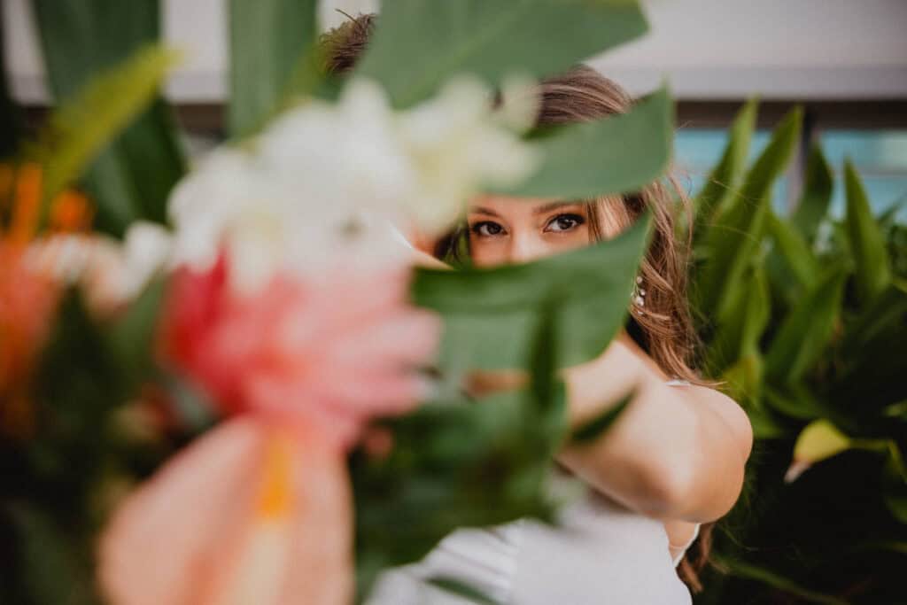 A woman partially obscured by a tropical plant holds a bouquet of flowers in front of her face, with only one eye and part of her hair visible through the foliage.