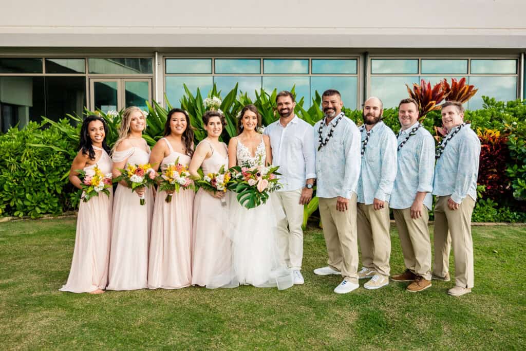 A wedding party stands outside in front of greenery, with four women in pink dresses on the left, the bride and groom in the center, and four men in light blue shirts and beige pants on the right.