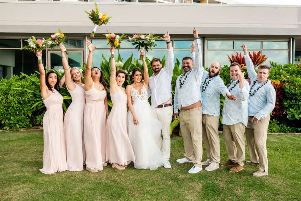 A newlywed couple stands with their bridal party, all cheering with raised arms and holding bouquets, in front of a green garden and colorful plants. The group is dressed in light, casual attire.