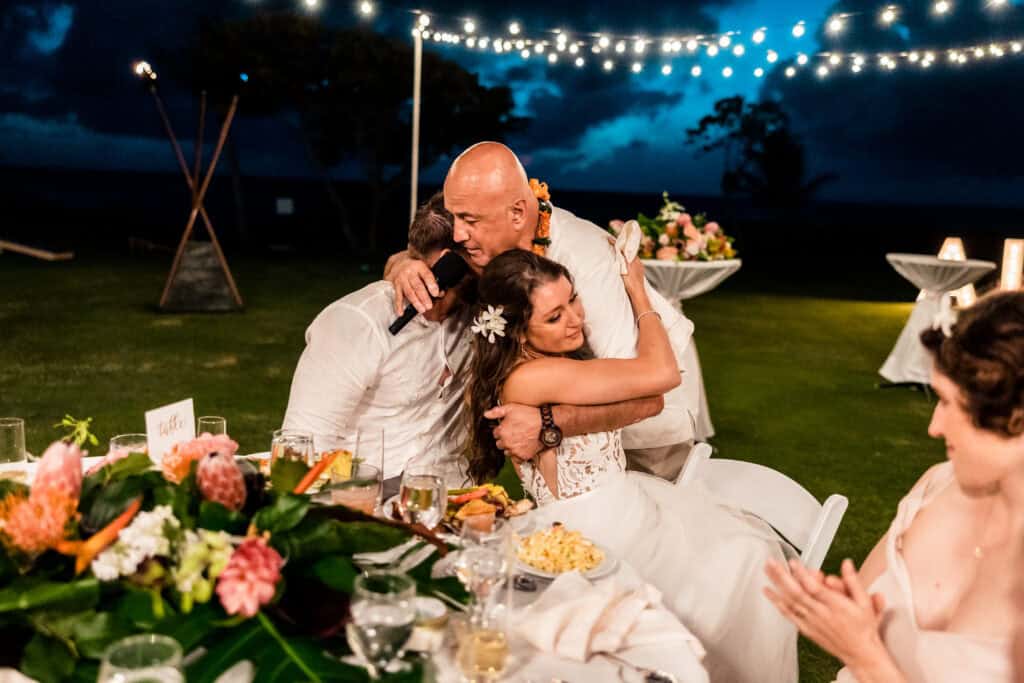 A bride and groom are embracing a man under string lights during an outdoor evening wedding reception. The tables are decorated with flowers, and guests are seated around them.