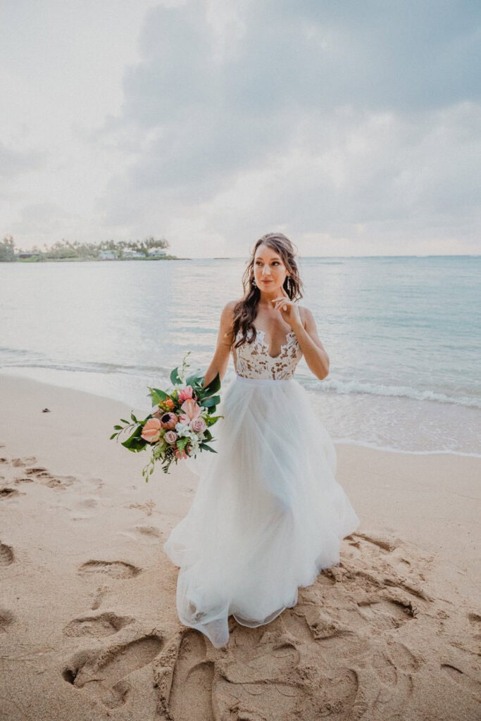 A woman in a white wedding dress stands on a sandy beach holding a bouquet of flowers, with the ocean and cloudy sky in the background.