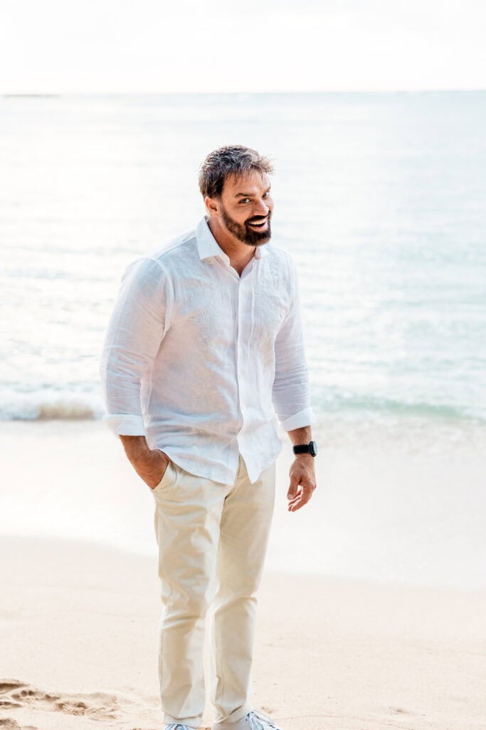 A man in a white shirt and beige pants stands on a sandy beach with the sea in the background, smiling.
