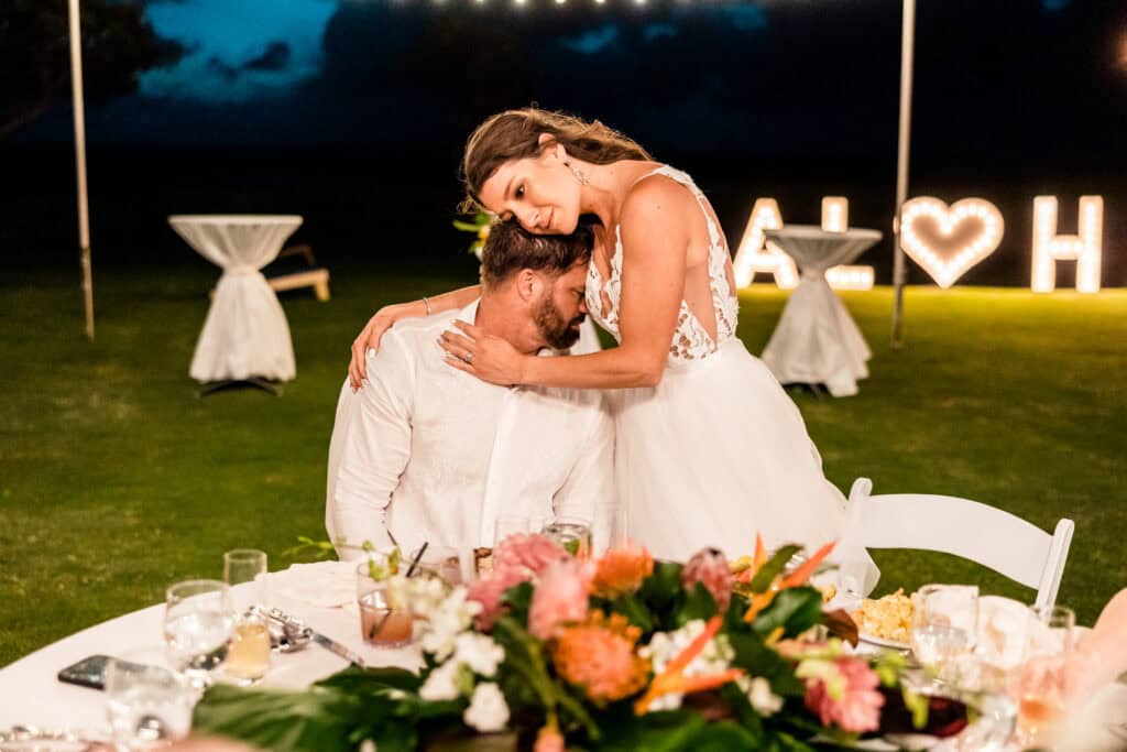 A bride stands beside a seated groom, resting her head on his, during an outdoor reception. A table with food is in the foreground, and large illuminated "A♥H" letters are in the background.