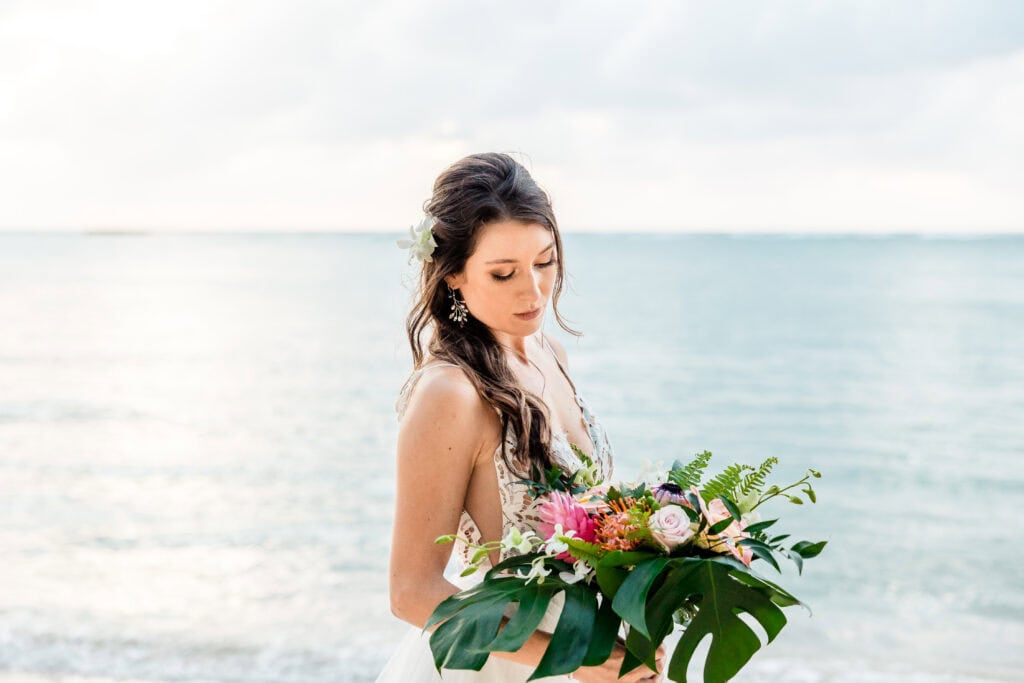 A woman in a light dress holding a bouquet of flowers stands on a beach with calm ocean waters in the background.