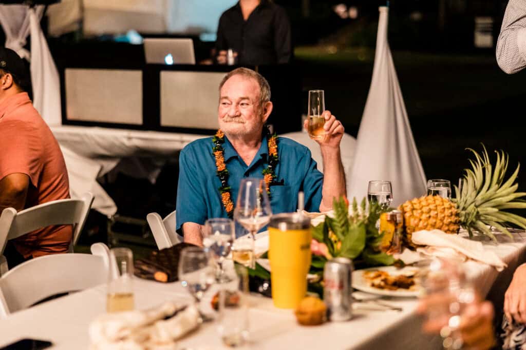 An older man in a blue shirt and lei raises a glass at a dinner event. He is seated at a table adorned with flowers, fruits, and drinks. Other guests are visible in the background.