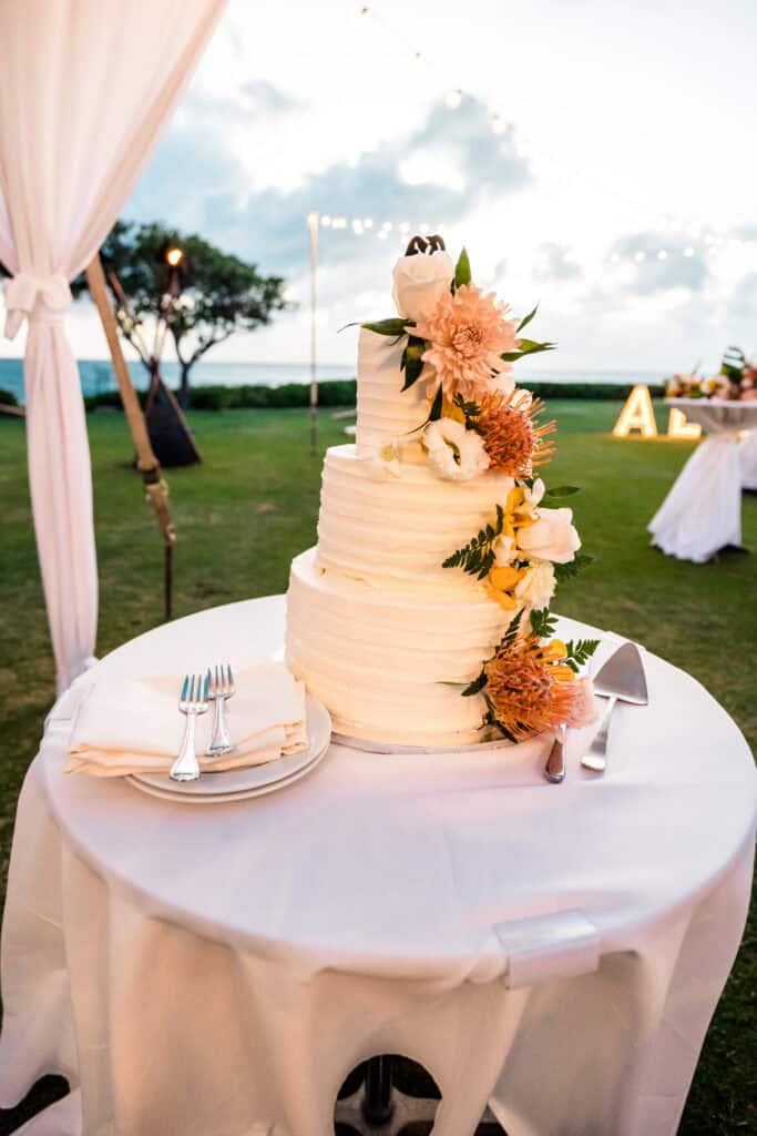 A three-tiered white wedding cake decorated with flowers stands on a round table covered with a white tablecloth; nearby are utensils and a cake server. White drapes and outdoor greenery are visible in the background.