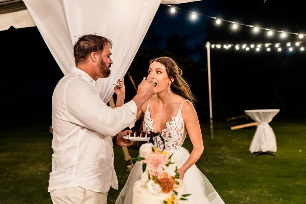 A groom feeds cake to a bride under a canopy with string lights overhead. The bride is in a white wedding dress, and the groom is in a white shirt. There is a tiered cake decorated with flowers in the foreground.