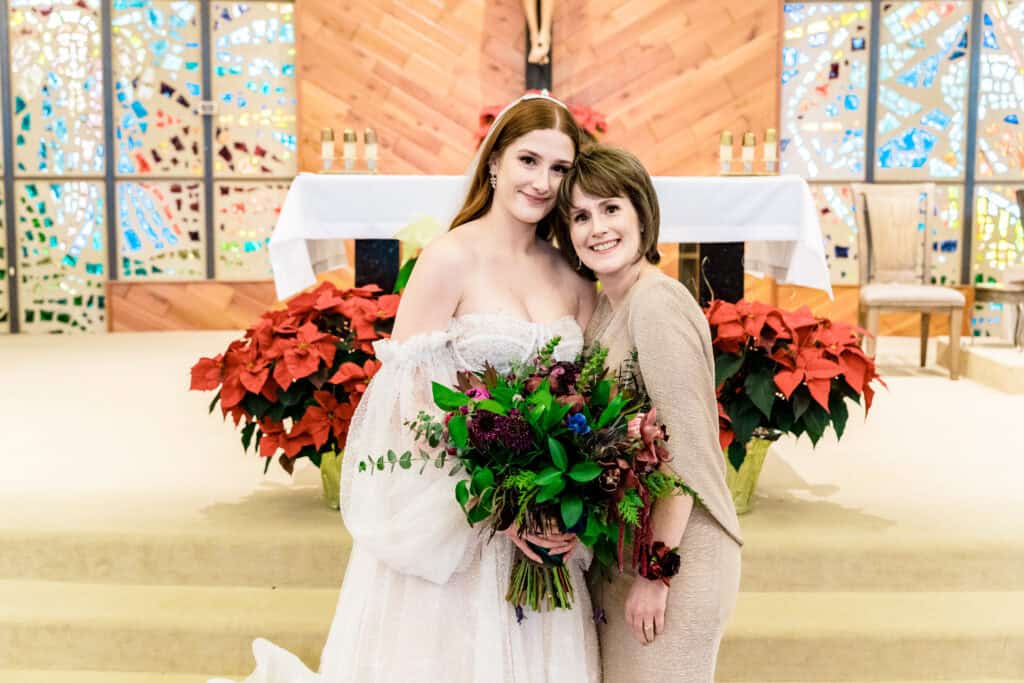 A bride in a white dress and a woman in a beige outfit stand closely together in front of an altar decorated with red flowers and stained glass windows.