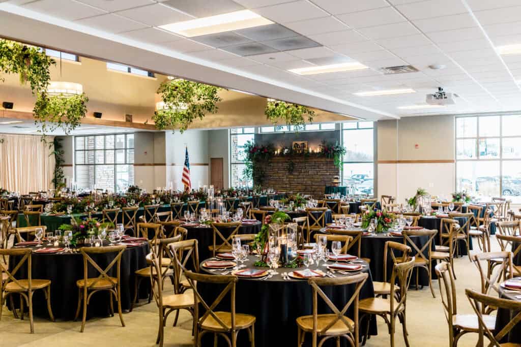 A large banquet hall set for an event with round tables, wooden chairs, floral centerpieces, and an American flag in the background.