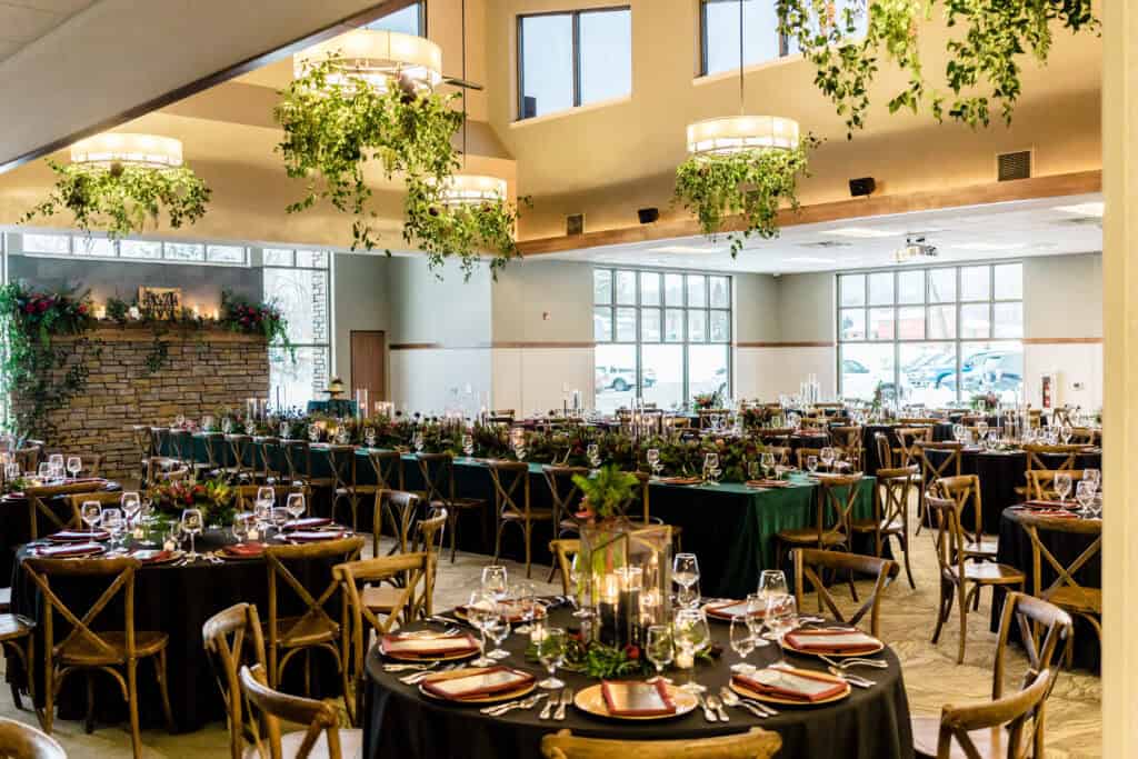 A banquet hall setup features multiple round tables with black tablecloths, floral centerpieces, and wooden chairs. Green and white hanging decorations are suspended from the ceiling.