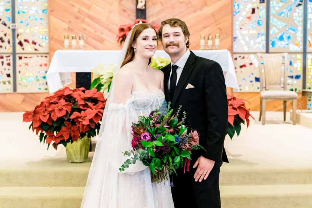 A bride and groom stand together in a church, smiling. The bride holds a bouquet of flowers and wears a white gown, while the groom is dressed in a black suit. Poinsettias and an altar are visible behind them.