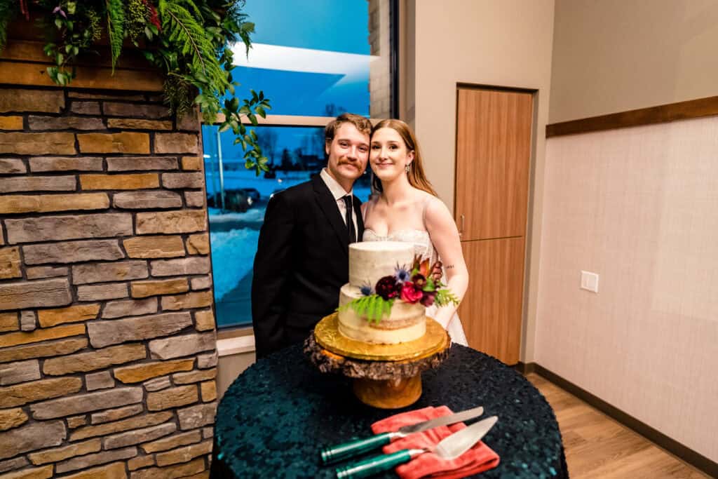 A bride and groom stand beside a decorated wedding cake on a table with a knife and server. They are inside a room with stone and wood decor, and there is snow visible outside the window.