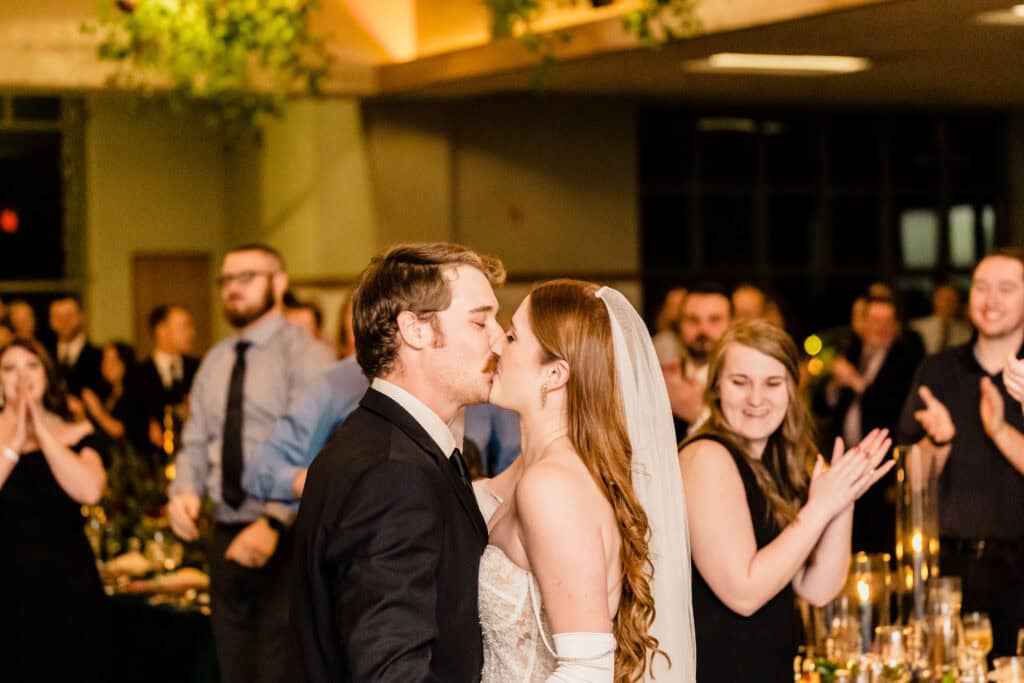 A bride and groom share a kiss at their wedding reception while guests stand and applaud in the background.