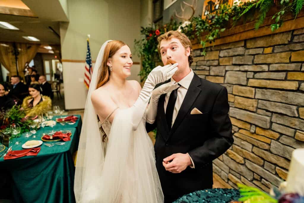 A bride in a white gown feeds her groom a piece of cake at a wedding reception. The groom is dressed in a black tuxedo. They are standing near a stone wall adorned with greenery.