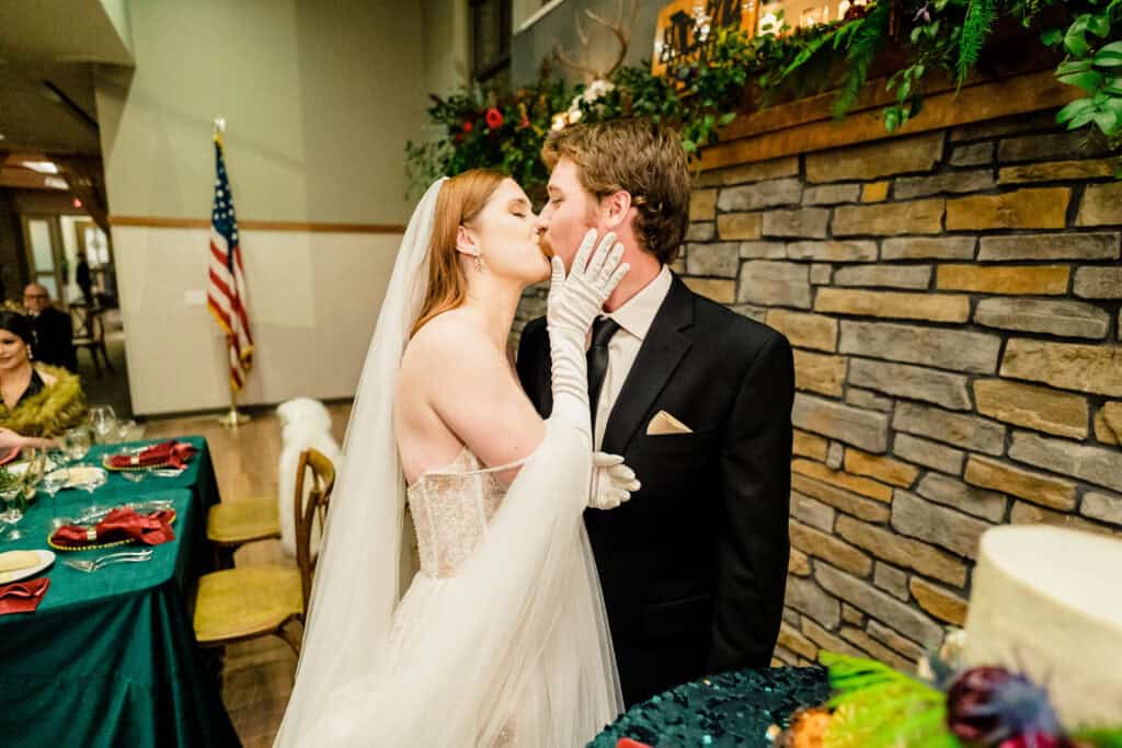 A bride in a white wedding dress and veil kisses a groom in a black suit and tie. They stand near a stone wall with a decorated table and an American flag in the background.