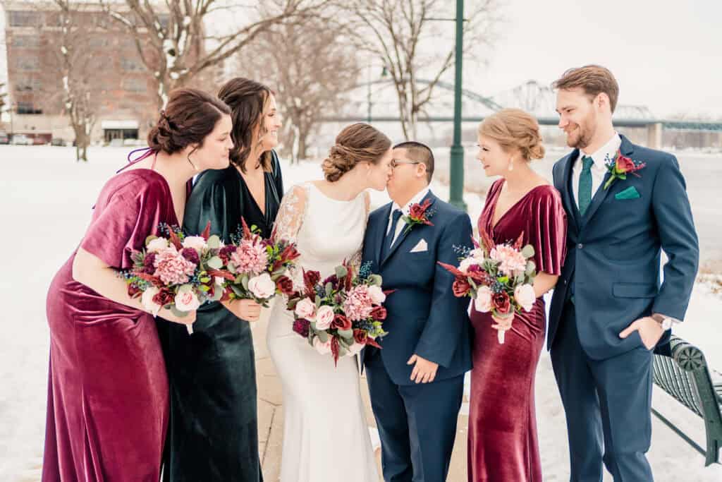 A wedding party stands outdoors in winter attire, smiling and holding bouquets. The group includes a bride in white, a groom in blue, two bridesmaids in velvet dresses, and one groomsman.