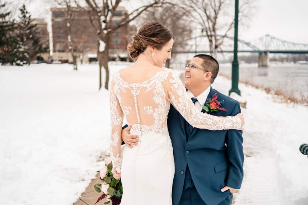 A couple, one in a white wedding dress with lace details and the other in a blue suit, embrace and smile at each other outdoors in a snowy setting with a bridge in the background.