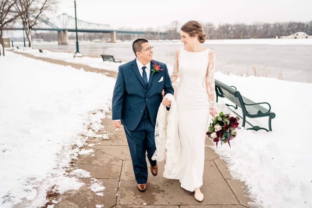 A couple walks hand in hand on a snowy path by a river, dressed formally in a navy suit and a white wedding dress, with a bridge visible in the background.