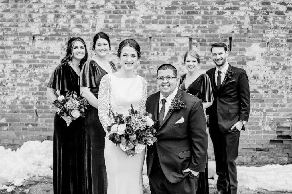 A bridal couple poses with four attendants in front of a brick wall. The bride holds a bouquet, and the group smiles. Snow is visible on the ground.