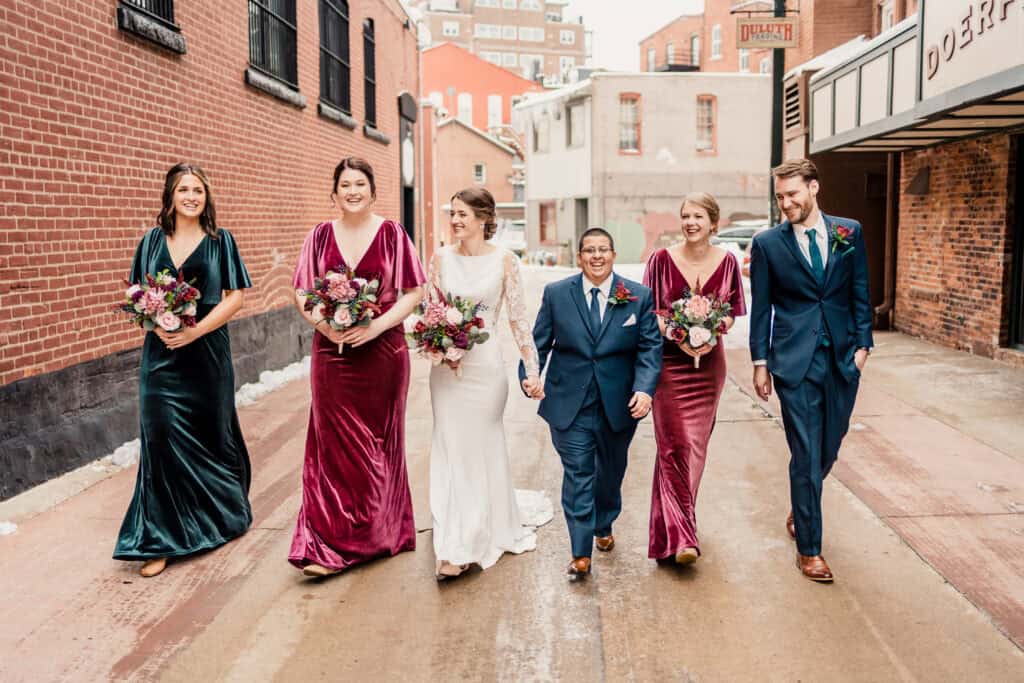 A wedding party walks down an alley. The group includes two people in wedding attire, with the rest in formal dresses and suits, all holding bouquets. They appear joyful and relaxed.