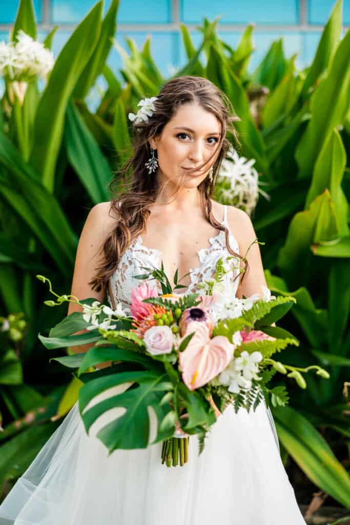 A bride with long, wavy hair holds a large floral bouquet featuring various green leaves and pink flowers. She stands in front of lush green plants and large leaves.