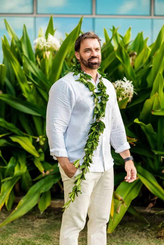 A man wearing a white shirt and beige pants stands in front of lush green plants. He has a leafy garland around his neck and shoulder.