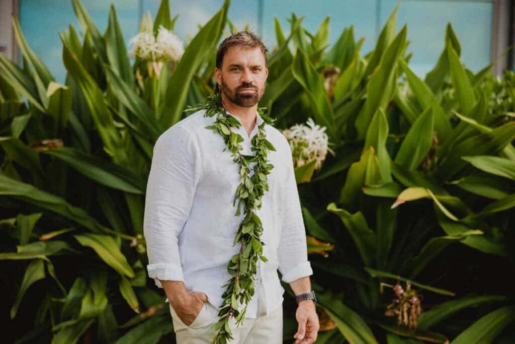 A man in a white shirt stands in front of lush green plants, wearing a leafy garland around his neck and wrist.