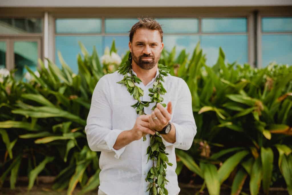 A man with a beard, wearing a white shirt and a leafy garland around his neck, stands in front of green foliage near a building.