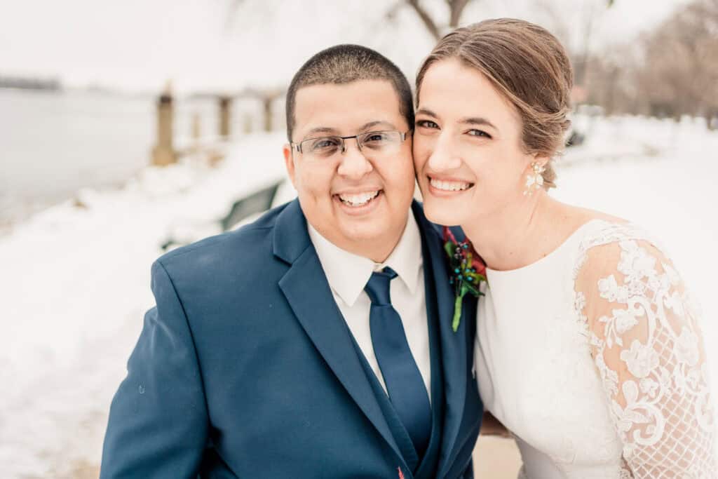 A smiling couple, one in a navy suit and the other in a white lace wedding dress, embrace outdoors on a snowy day with a waterfront in the background.