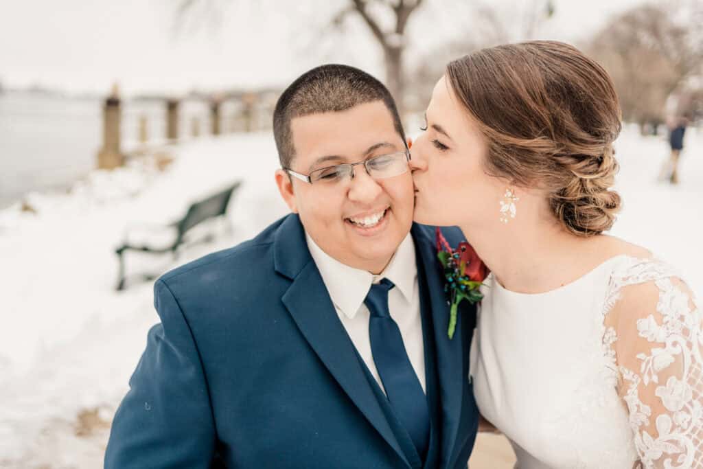 A bride in a white dress and groom in a blue suit are smiling as she kisses his cheek in a snowy outdoor setting.