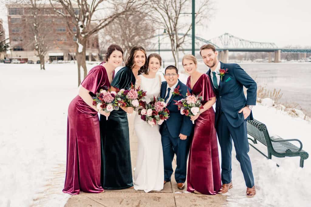 A group of five people dressed in formal attire pose together outdoors on a snowy day, three holding flower bouquets. A bridge and trees are visible in the background.