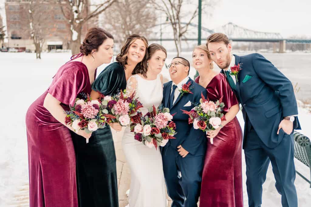 A wedding party poses outdoors in winter attire. The bride and groom are surrounded by bridesmaids in velvet dresses and groomsmen in suits, all holding bouquets of flowers. A bridge is visible in the background.