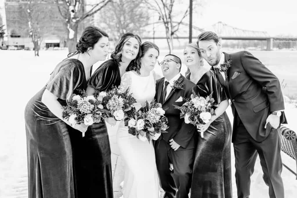 A wedding party, consisting of three bridesmaids holding bouquets, two groomsmen, and the bride in the center, poses cheerfully outdoors in a snowy setting. A bridge is visible in the background.