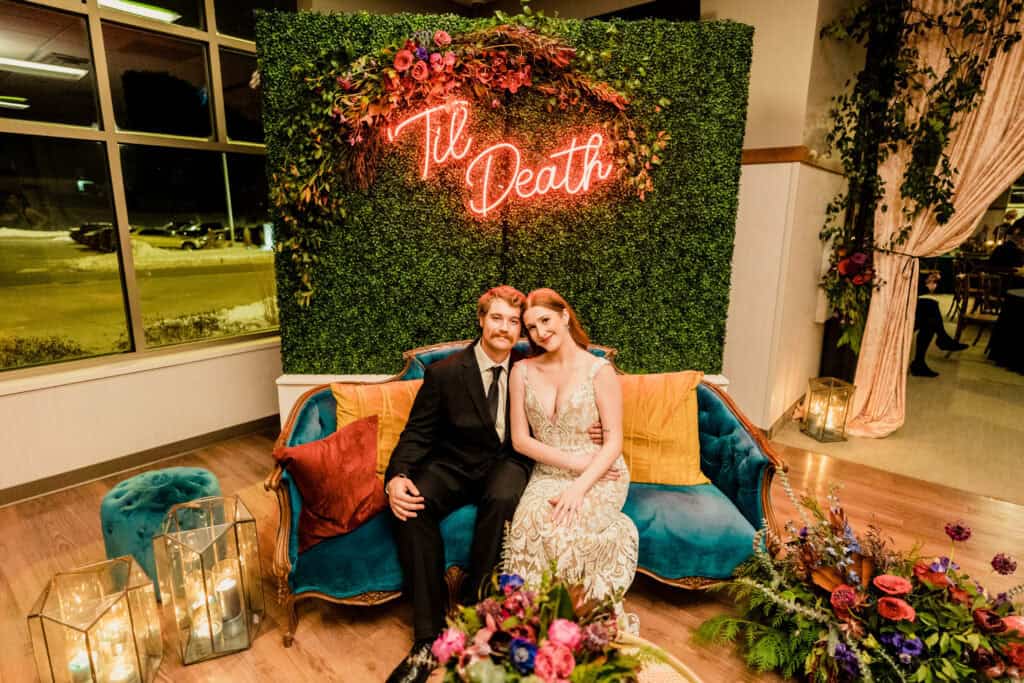 A couple dressed formally sits on a blue couch, with a neon sign reading "Til Death" above them and floral decorations surrounding the area.