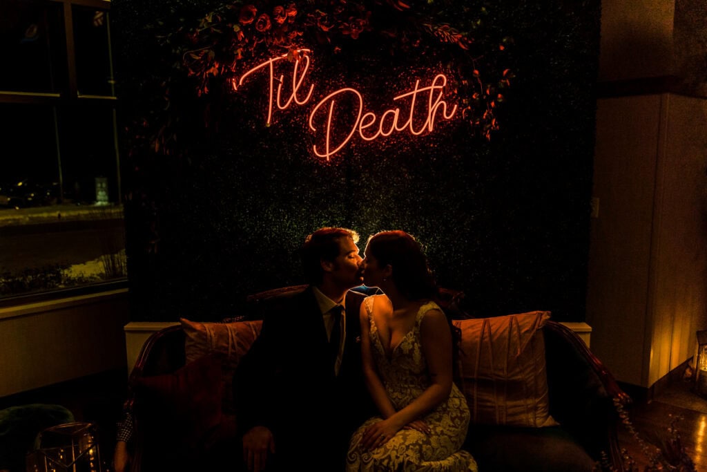 A couple sits closely together on a couch, kissing under a neon sign that says "Til Death." The background is dark, and the scene is dimly lit, creating a romantic atmosphere.