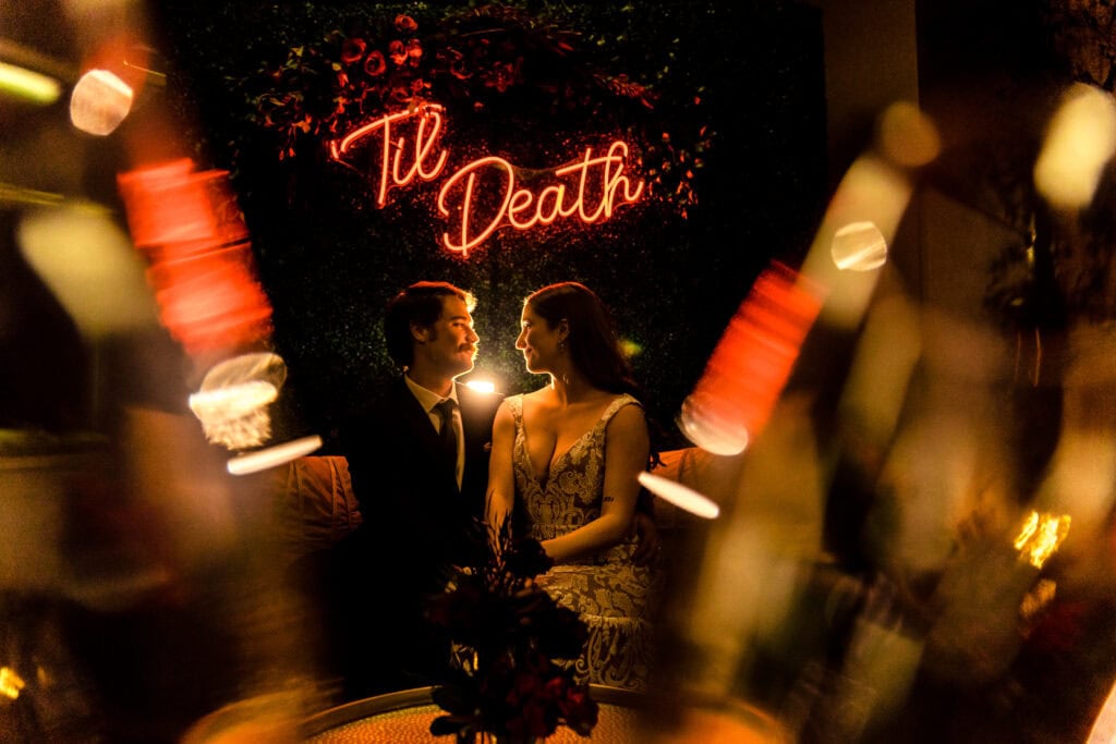 A couple sits closely and gazes at each other in a dimly lit room. Behind them, a neon sign reads "Til Death.