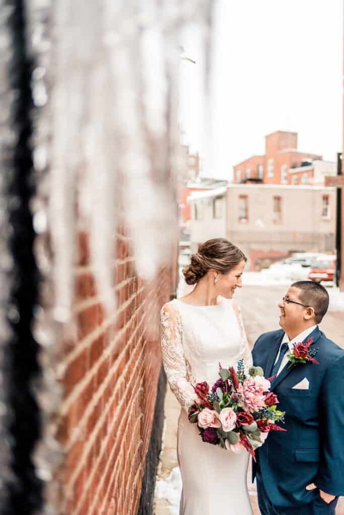 A couple stands outside near a brick wall on a snowy day. One person is wearing a white dress and holding a bouquet, while the other is dressed in a dark suit.