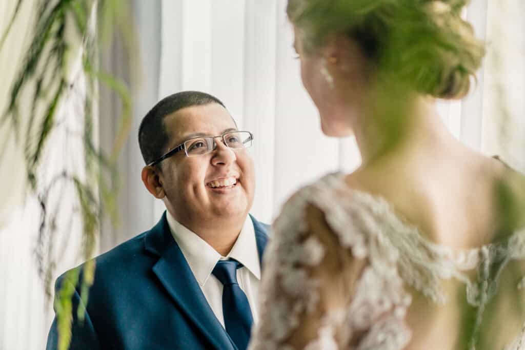 A man in a blue suit and glasses gazes and smiles at a woman in a white lace dress, partially obscured by foreground foliage.