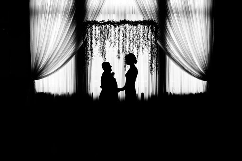 Silhouette of a man and woman facing each other, holding hands in front of a large window with sheer curtains and a decorative floral arch overhead.